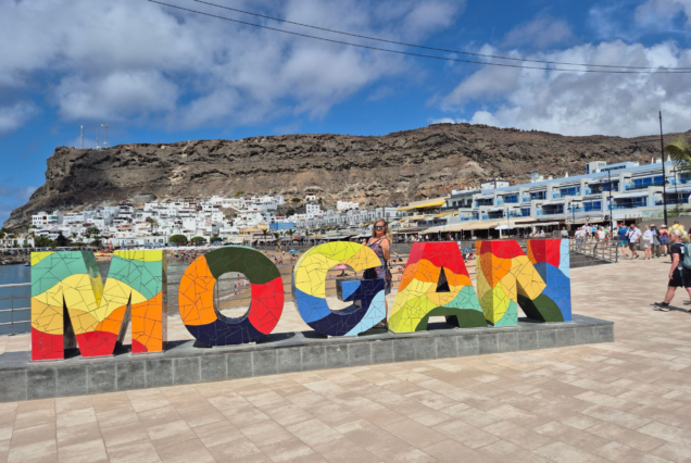 Cartel colorido que dice "MOGÁN" en el puerto de Mogán, con fondo de casas blancas y montañas, ideal para un viaje de avistamiento de delfines.
