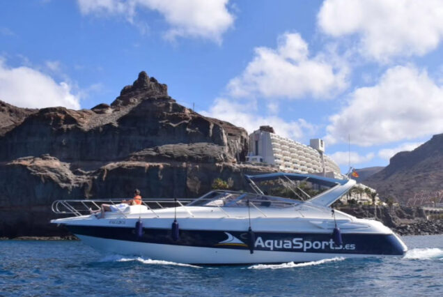 Yate de AquaSports navegando en aguas cristalinas frente a un paisaje montañoso y un hotel en la costa, bajo un cielo azul con nubes.